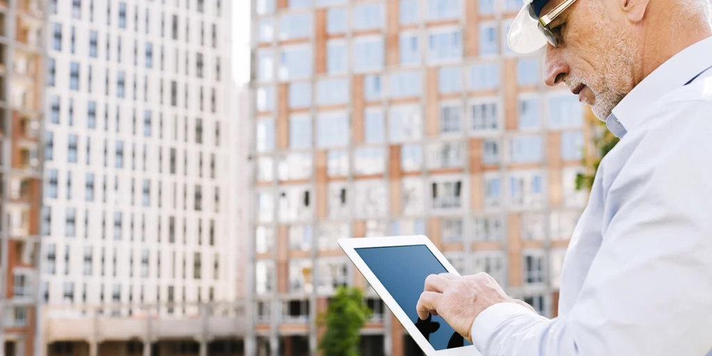 A imagem apresenta um homem com um tablet na mão, posicionado em frente a dois prédios. 