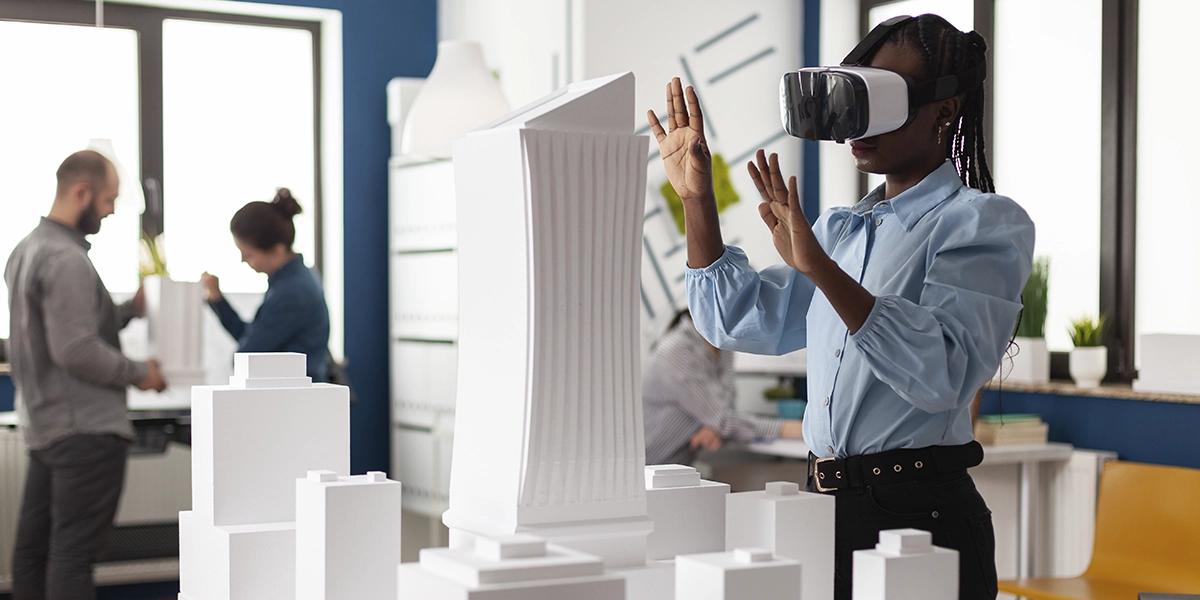 A imagem apresenta três pessoas em uma sala, sendo duas mulheres e um homem. Uma das mulheres está usando um óculos de realidade virtual, posicionada em frente a maquetes.