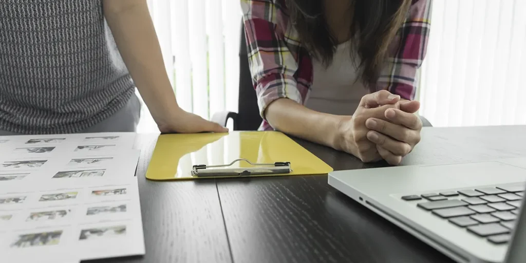 A imagem apresenta duas mulheres em uma mesa, mostrando apenas seus troncos e mãos. Na mesa, podemos ver um notebook e algumas folhas com imagens. 