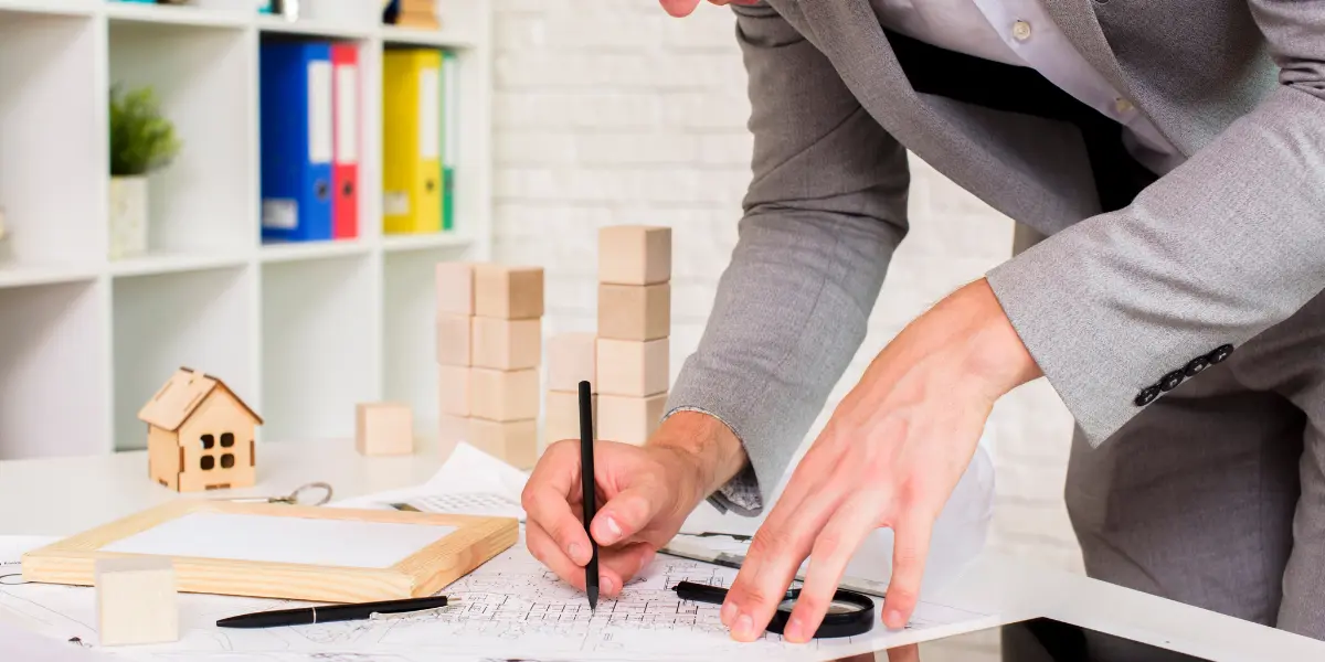 Gestão de projetos imobiliários: na imagem, um homem com terno cinza assinando um documento. Na mesa vemos alguns objetos de madeira como uma casa e alguns blocos