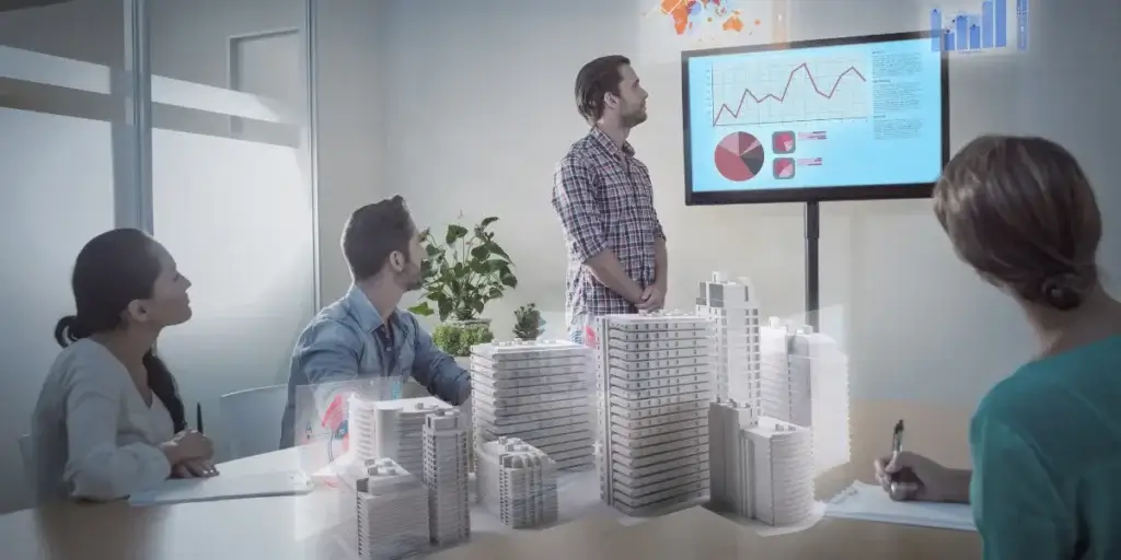 Business Intelligence no mercado imobiliário: Imagem mostra um homem em pé, ao lado de uma tela com gráficos. Ao redor de uma mesa há uma mulher e um homem sentados lado a lado, e uma outra mulher também sentada em frente a eles. Na mesa há a projeção de imagem 3D de prédios. Há um vaso de planta no canto da sala onde todos estão. 