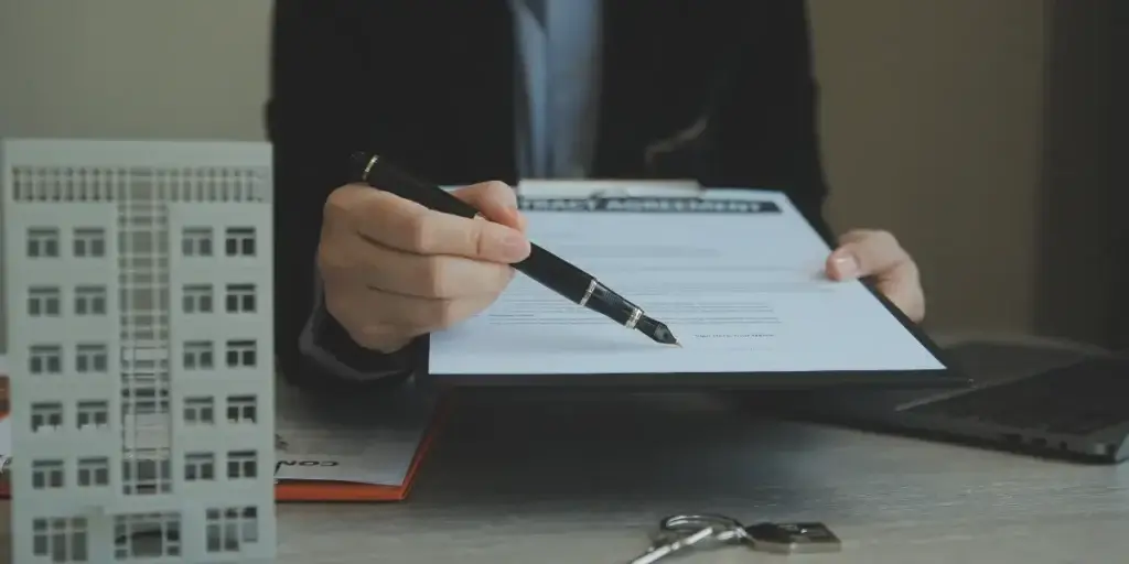 Lavratura de escritura Lavratura de escritura: Imagem mostra o tronco de homem segurando uma prancheta com um documento. Na mão ele segura uma caneta apontando para a assinatura. Há uma miniatura de um prédio. e uma chave.