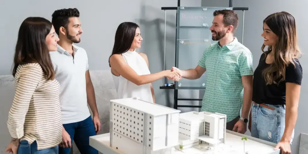 Plantão de vendas Plantão de vendas: Cinco pessoas, sendo três mulheres e dois homens, estão em volta de uma mesa onde há uma maquete realista de um prédio. Um homem e uma mulher apertam as mãos.