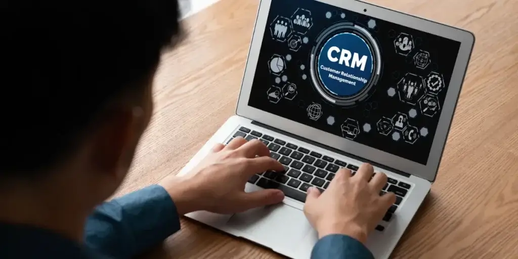 CRM de alta performance CRM de alta performance: Imagem mostra um homem sentado, com um notebook em frente a ele, em cima de uma mesa, onde ele está digitando. Na tela há ícones relacionados a tecnologia e o símbolo do CRM.