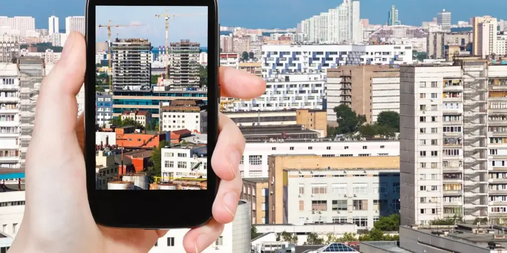 conteúdo para o mercado imobiliário conteúdo para o mercado imobiliário: Imagem mostra uma mão segurando um smartphone, capturando a imagem de prédios em uma cidade.