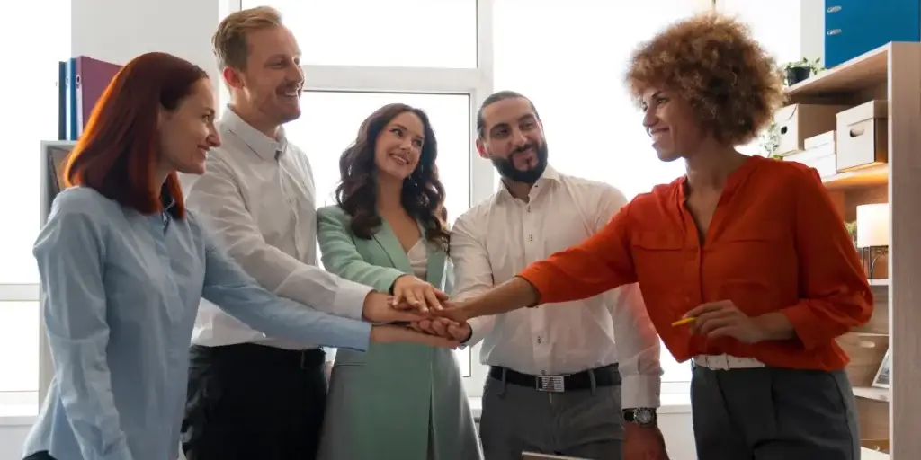 CRM para equipe de vendas imobiliárias CRM para equipe de vendas imobiliárias: imagem de 5 pessoas num escritório, com as mãos juntas no centro de uma roda. Eles se olham e sorriem.