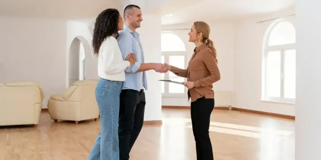 omnicanalidade no mercado imobiliário omnicanalidade no mercado imobiliário: imagem mostra um homem e uma mulher lado a lado, ela segurando no braço dele. Em frente a eles há uma outra mulher, dando aperto de mãos no homem e segurando uma prancheta, Eles estão em uma sala de uma casa, há janelas e duas poltronas atrás deles.