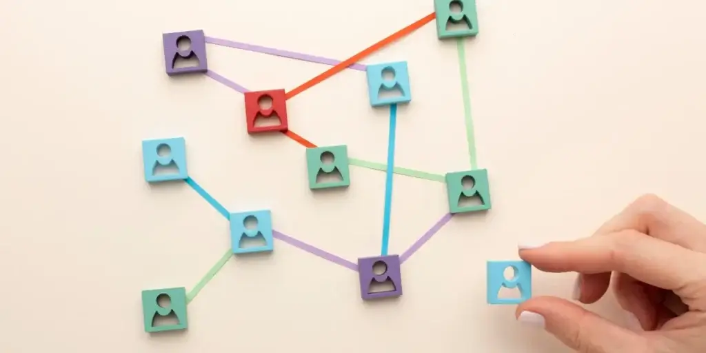 jornada do cliente imobiliário: A composição centraliza-se em uma rede complexa de blocos quadrados de várias cores, cada um contendo uma silhueta simples de uma figura humana. As linhas finas e coloridas conectam os blocos, formando uma rede que sugere conexões ou interações. Uma mão feminina, com unhas pintadas, posiciona-se para adicionar um bloco azul, o que enfatiza a ação de construção ou a criação de uma nova ligação dentro da rede.