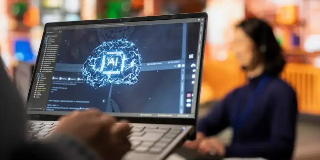 CRM com IA CRM com IA: Imagem mostra um notebook em primeiro plano, com a mão de uma pessoa no teclado, e na tela a imagem tecnológico de um cérebro com as iniciais IA em cima. Em segundo plano há uma mulher sentada.