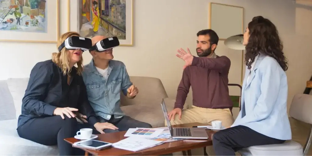 Tour virtual imóvel: A composição da cena é organizada de forma que os dois indivíduos com óculos de RV estão sentados em um sofá, no centro, enquanto um homem e uma mulher estão posicionados em frente a eles, ao redor de uma mesa de centro. O homem, gesticulando com as mãos, parece estar explicando algo. A mulher, sentada e olhando para a dupla com os óculos, está envolvida na conversa. A luz entra pela janela, iluminando a cena e os elementos.