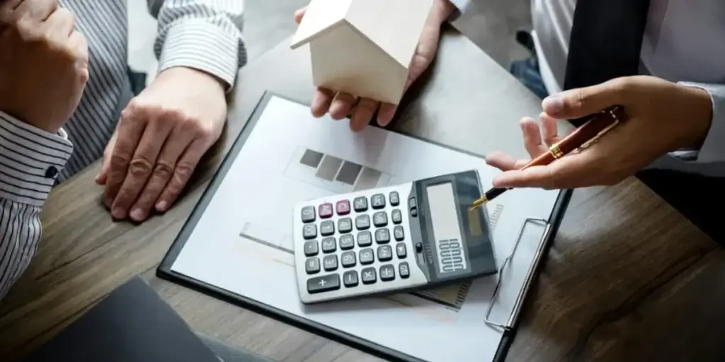 Comissão imobiliária Comissão imobiliária: imagem mostra os braços de dois homens que estão sentados. Um deles segura uma caneta em uma das mãos e uma miniatura de casa na outra. No centro da imagem, em uma mesa, há uma prancheta com uma calculadora em cima.