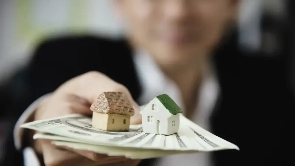 gestão de carteira imobiliária gestão de carteira imobiliária: um homem em segundo plano, com a imagem desfocada, segura um maço de dinheiro com uma das mãos, que está apontada para frente. Em cima do dinheiro há duas miniaturas de casas.