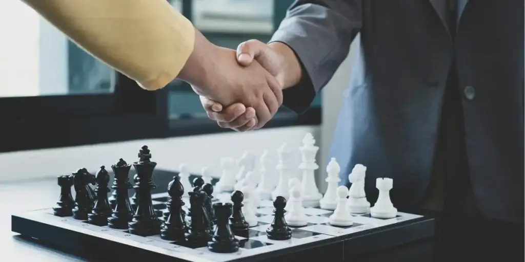 Treinamento de corretores: imagem mostra os braços de duas pessoas que estão se cumprimentando dando um aperto de mãos. Embaixo desta cena há um jogo de xadrez. 