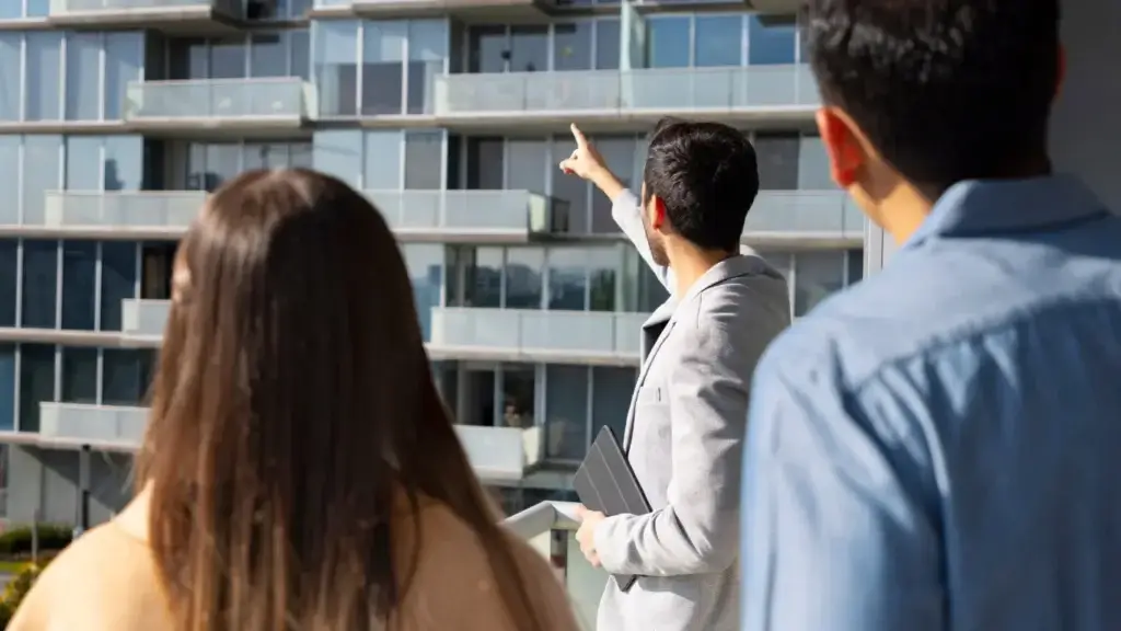 omnichannel imobiliário: Três pessoas observam um prédio residencial moderno com varandas envidraçadas, enquanto um homem aponta para um dos apartamentos, indicando interesse ou explicação sobre o imóvel.