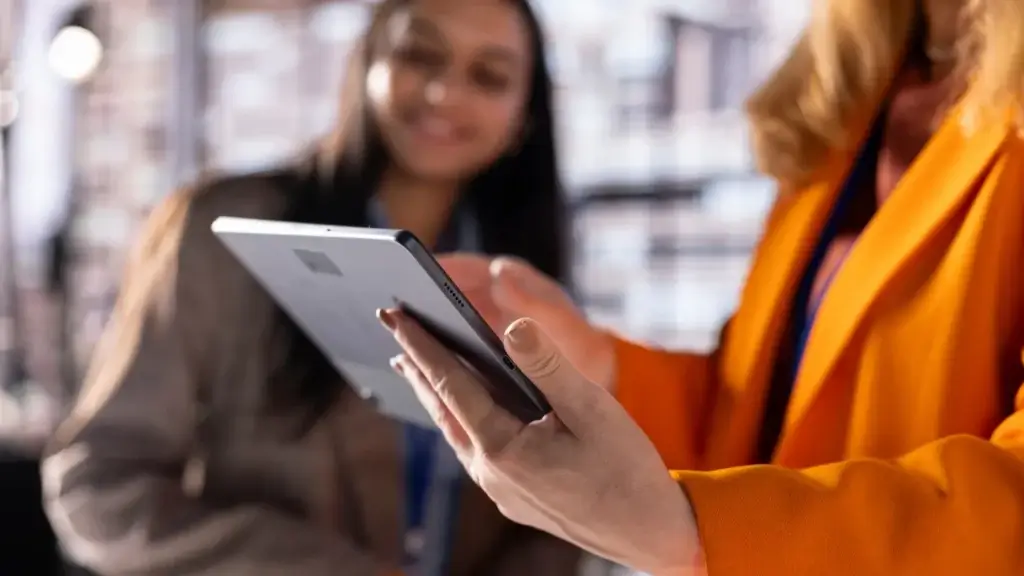 omnichannel imobiliário: Pessoa segurando um tablet e mostrando a tela para outra pessoa ao fundo, em um ambiente interno corporativo, sugerindo análise ou apresentação de informações digitais.