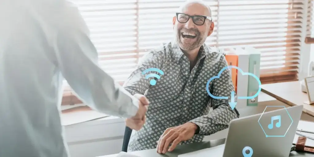 Script de vendas pelo WhatsApp imobiliário: A cena é composta com foco no aperto de mão entre dois indivíduos, que ocupam a parte central e direita da imagem. O homem à direita está sentado em uma mesa, olhando para a frente e sorrindo amplamente, enquanto o outro homem, que mostra apenas as costas, de pé e de costas para a câmera, estende a mão para cumprimentá-lo. Há um notebook na mesa, livros empilhados e uma cortina persiana na janela ao fundo da imagem. 