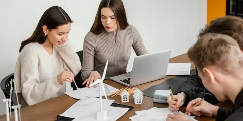Antecipação de repasse imobiliário: em volta de uma mesa de madeira há um grupo de jovens adultos, composto por duas mulheres e dois homens. Uma mulher à esquerda, com cabelo escuro e longo, usa um suéter branco creme e está curvada sobre papéis, segurando uma caneta. Ao seu lado, uma mulher com cabelo castanho longo e liso, vestindo um suéter de gola alta bege, olha para a tela de um laptop aberto à sua frente. Na frente, dois homens, um com cabelo castanho claro visível de costas, e outro mais ao fundo com cabelo escuro e ondulado, estão ambos concentrados em papéis e escrevendo. Na mesa, há modelos de casas e turbinas eólicas.
