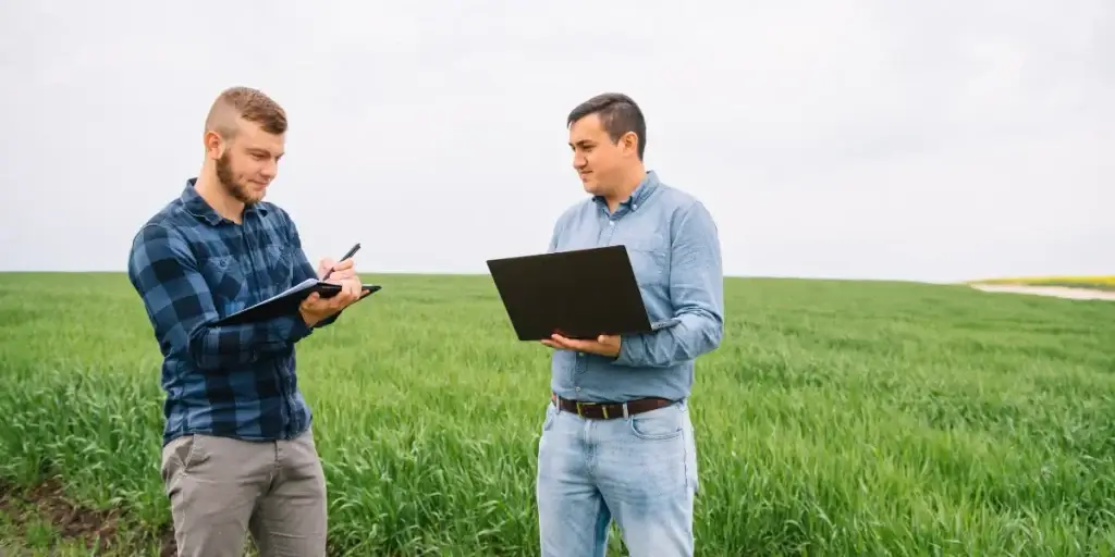 Como vender lotes encalhados: imagem mostra dois homens em pé, um de frente para o outro, em um terreno vazio, somente com grama. Um dos homens segura um notebook e o outro segura um caderno com uma caneta. 
