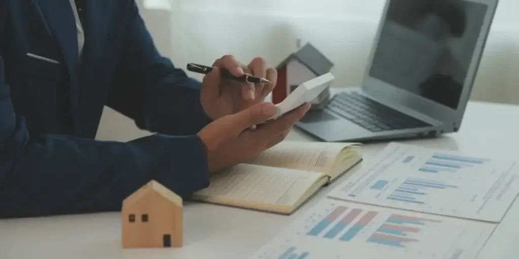 Estratégia de gestão de lançamentos imobiliários Estratégia de gestão de lançamentos imobiliários: A composição da cena é focada em uma mesa de escritório, com um corte próximo do tronco para cima de um indivíduo. A ação principal é a manipulação de uma calculadora, com uma caneta na outra mão. Na mesa há um laptop aberto, gráficos impressos em papel espalhados e um caderno de anotações, além de uma miniatura de casa feita em madeira.
