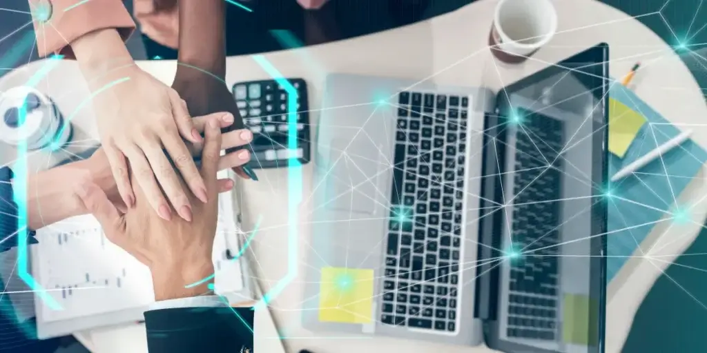 Gestão digital: A composição da imagem é um plano aéreo de uma mesa de escritório, com um foco central nas mãos de várias pessoas empilhadas umas sobre as outras em um gesto de unidade. Elementos de escritório como laptops, calculadoras e cadernos estão arranjados ao redor, sugerindo um ambiente de trabalho dinâmico. linhas digitais, em forma de uma rede abstrata, cruzam a cena, conectando visualmente os diferentes componentes.