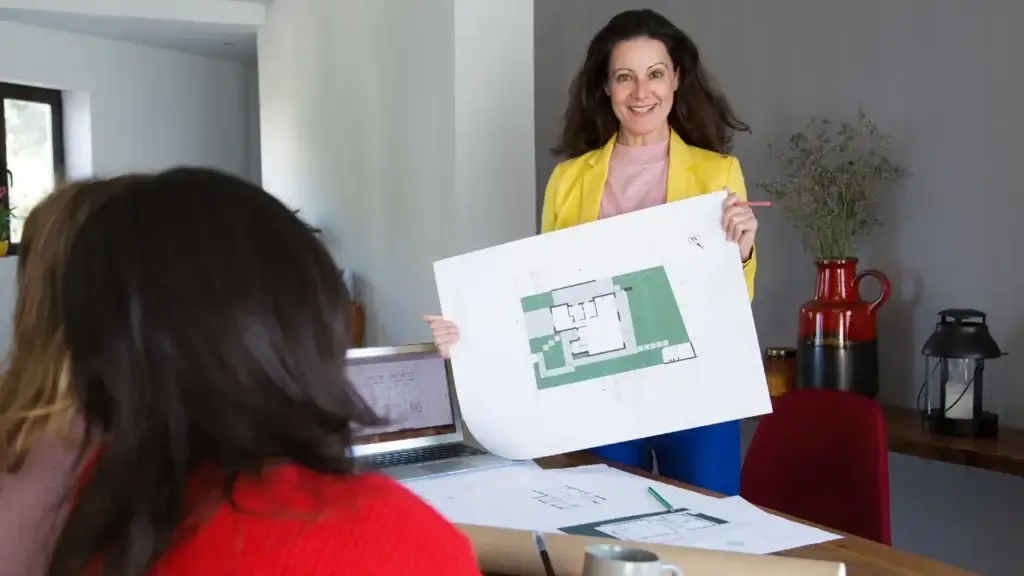 gestão para loteadoras: A composição da cena é focada em uma interação de trabalho, com uma mulher posicionada centralmente atrás de uma mesa, servindo como o ponto focal. Duas outras mulheres estão sentadas à frente, vistas de costas, o que direciona o olhar do espectador para o plano arquitetônico exibido. A disposição dos elementos, incluindo um laptop aberto e plantas espalhadas sobre a mesa de madeira, cria uma atmosfera realista de consulta técnica e planejamento residencial. A profundidade da cena é bem definida pela posição dos sujeitos e pelo mobiliário ao fundo, com uma mesa, um jarro com planta e uma luminária.