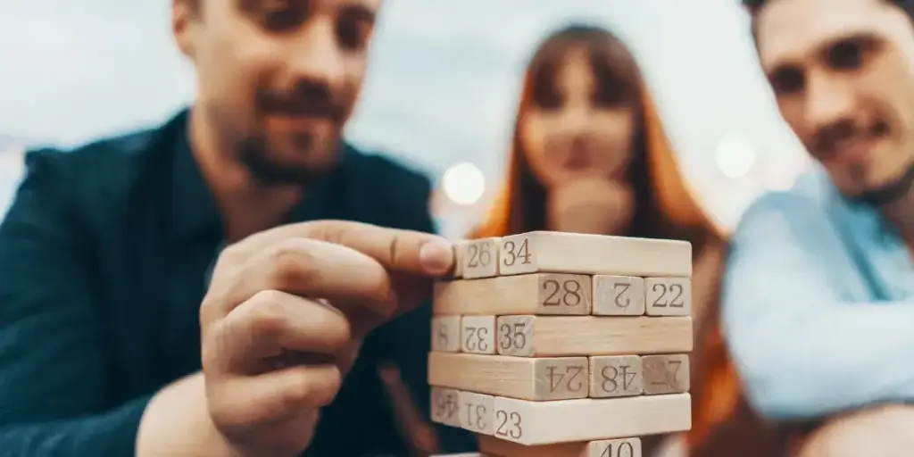 implantar CRM imobiliário implantar CRM imobiliário: Uma fotografia em close-up captura um momento de concentração enquanto um grupo de dois homens e uma mulher, desfocados, joga uma versão numérica de um jogo de empilhamento de blocos de madeira.