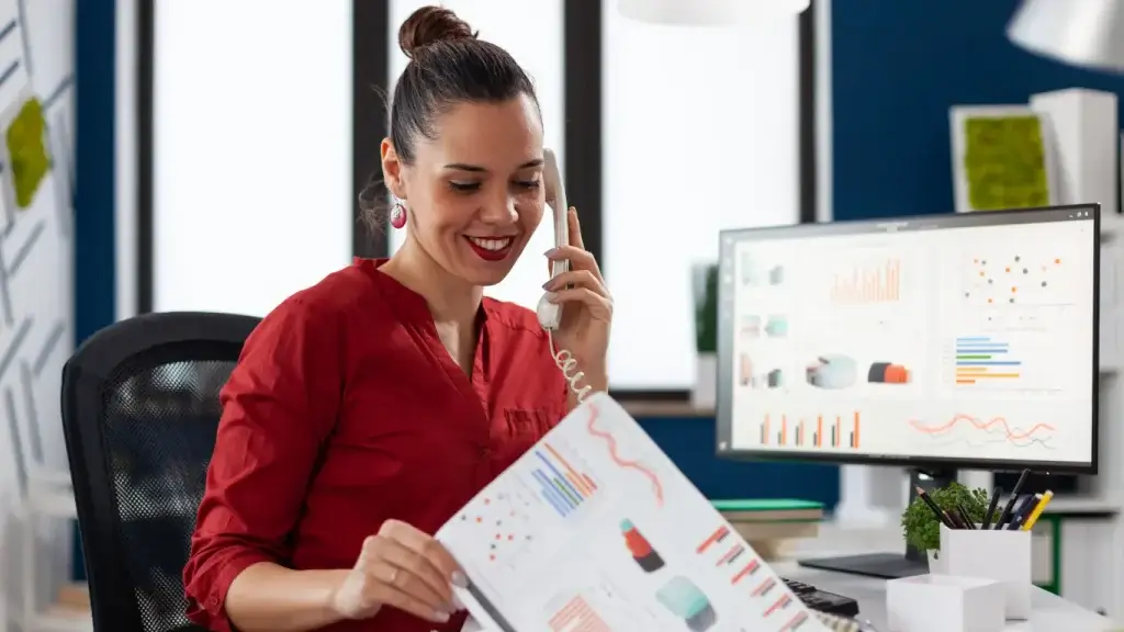 accountability comercial accountability comercial: uma mulher sentada em frente a uma tela de computador, onde há vários gráficos. Em uma das mãos ela está cum um telefone fixo e na outra segura uma folha de papel, também com gráficos. Na mesa há um porta lápis. No fundo a imagem distorcida mostra uma janela grande, de vidro.