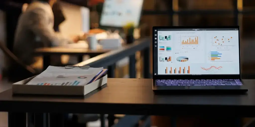 Nova era do CRM 8.0: imagem mostra um notebook aberto, com gráficos na tela. Ao lado dele tem uma prancheta com folhas com gráficos. Ao fundi da imagem, distorcida, tem uma pessoa sentada em frente a um computador.
